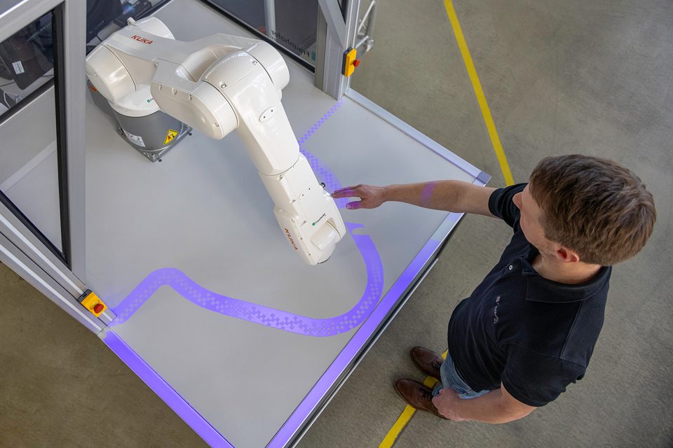 Person beside a robotic arm with a projected blue safety boundary on the work surface.