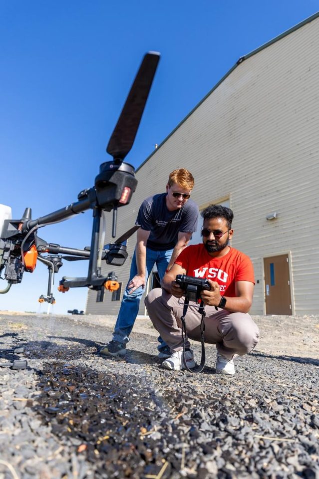 Two WSU researchers inspect a handheld controller beside a large agricultural spray drone.